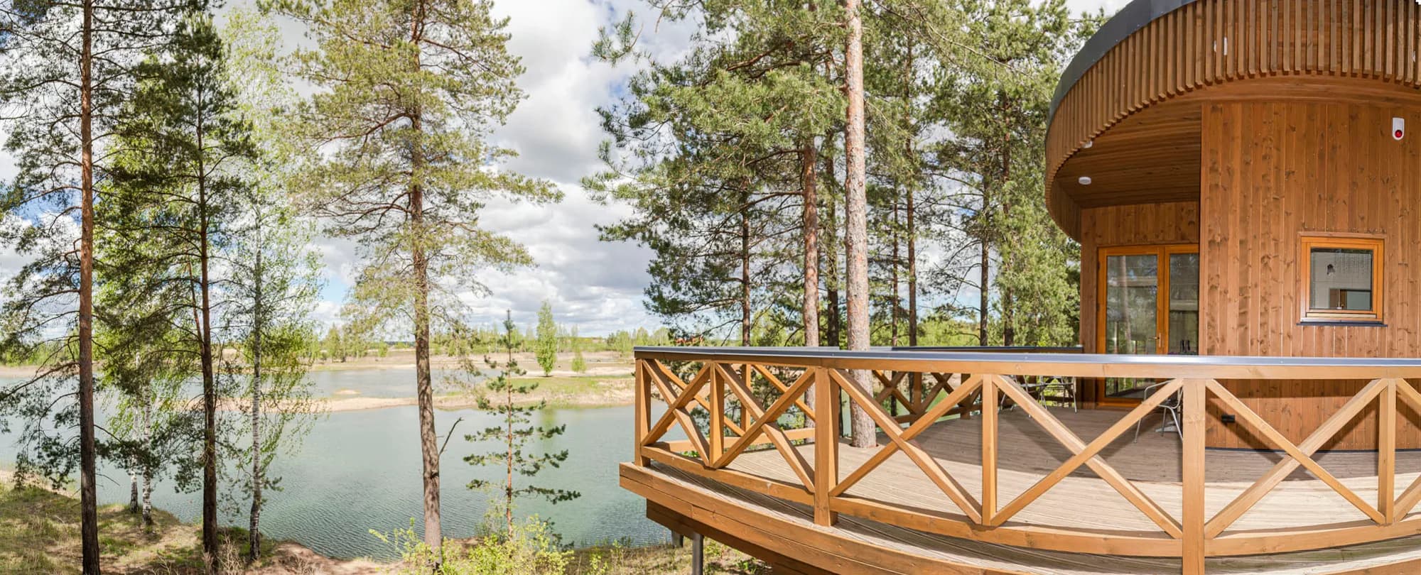 Panorāmas foto apaļai koka terasei pie ezera māju, ko ieskauj priedes un bērzi
